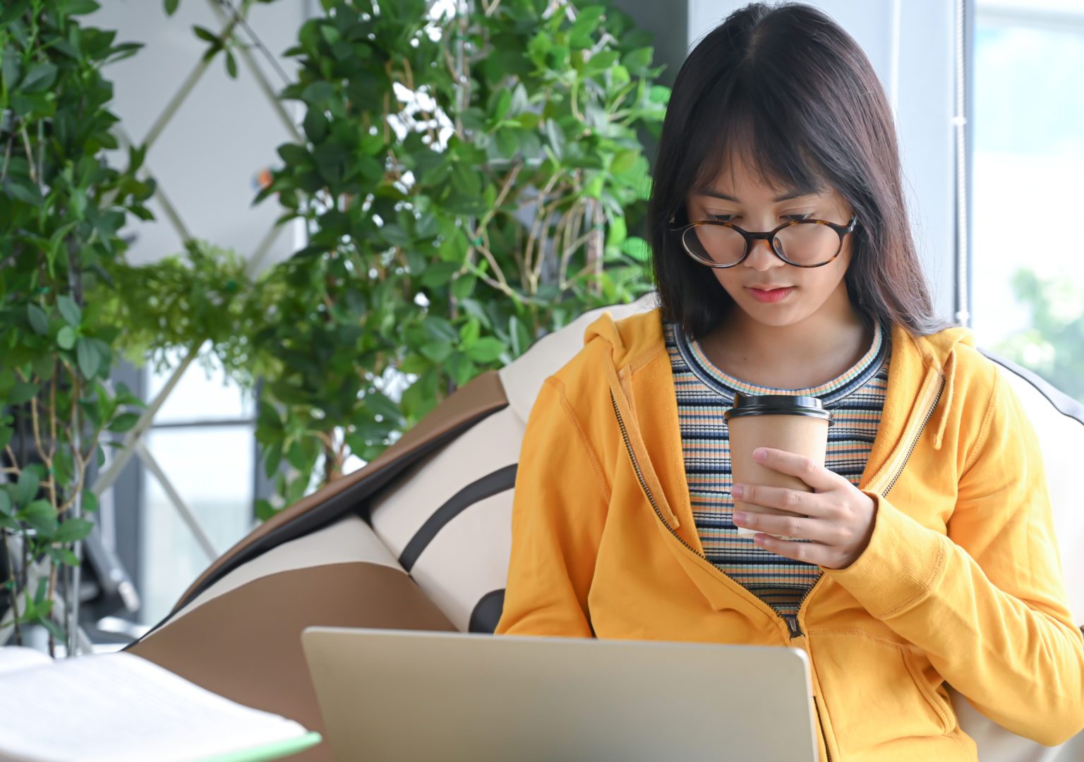 Asian female student wearing glasses is researching with a laptop to report, the other hand holding a coffee cup.