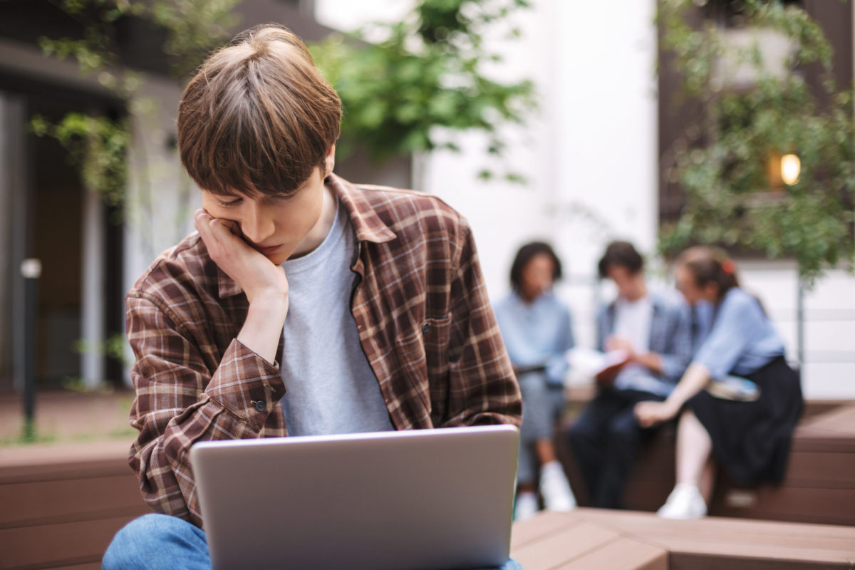 Photo of young tired man sitting on bench with laptop on knees and thinking while spending time in courtyard of university with students on background