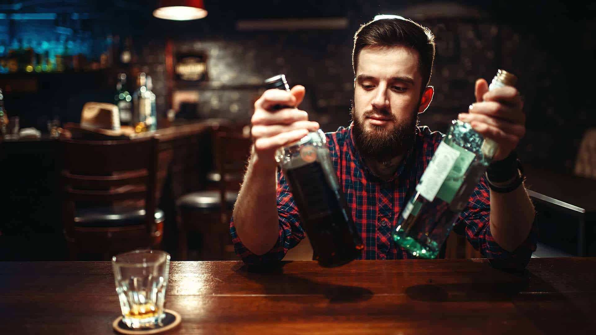 Alcoholic sitting at the bar counter with two bottles of alcohol beverage in hands. Male person in pub, alcoholism, drunkenness