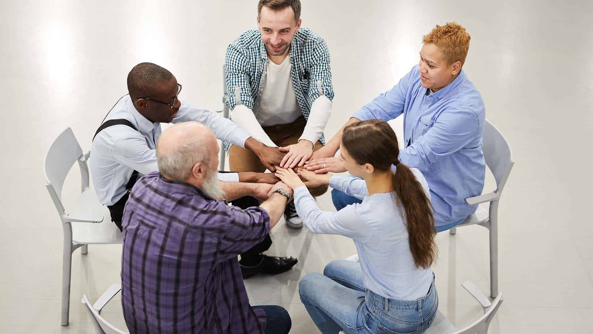High angle portrait of people joining hands sitting in circle during therapy session in support group, copy space