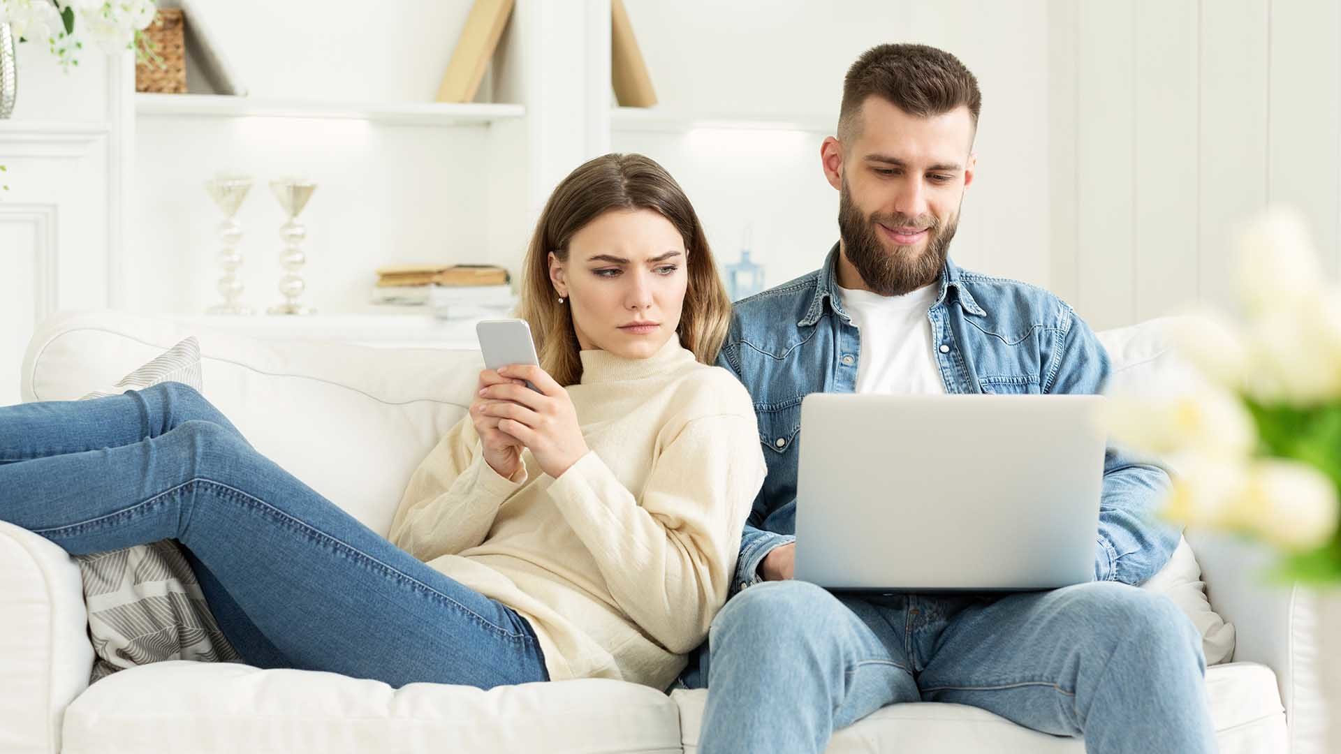 Woman looking at husband's laptop, sitting together on sofa with cellphone