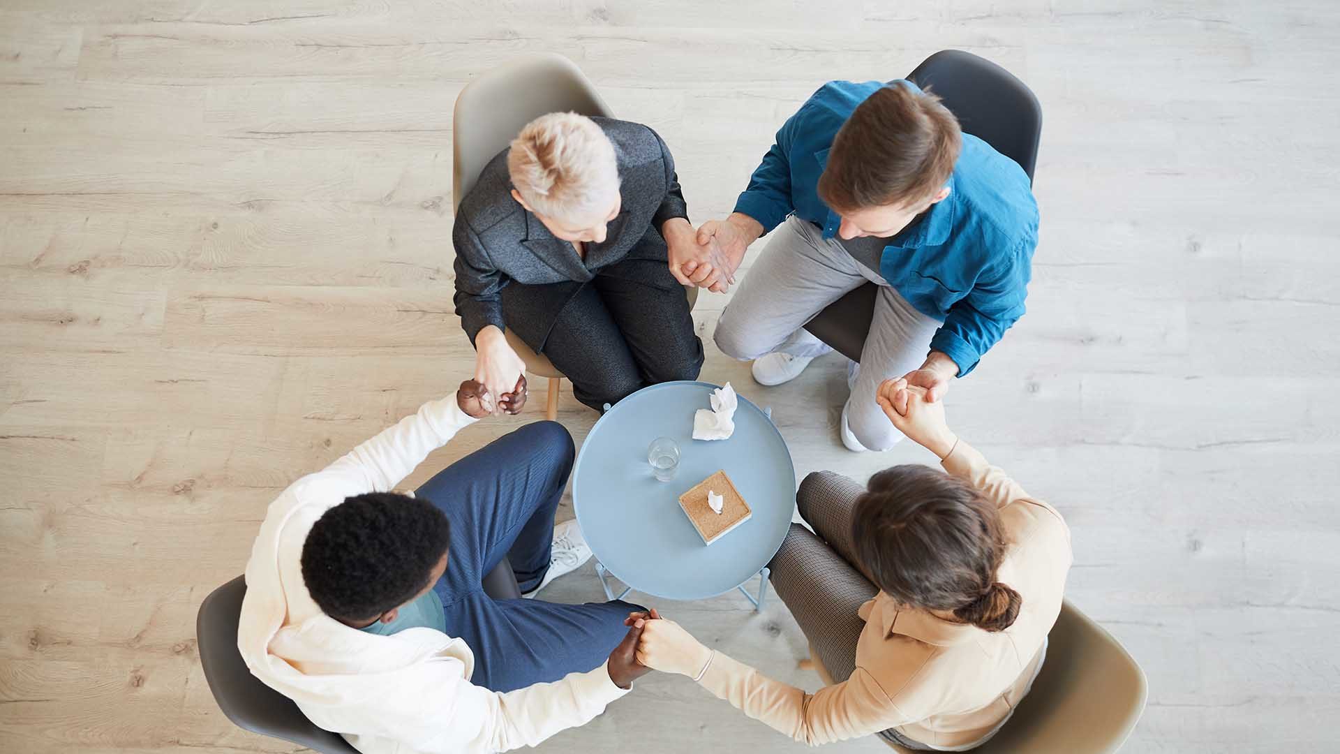 Above view at multi-ethnic group of people holding hands sitting in circle during therapy session in support group