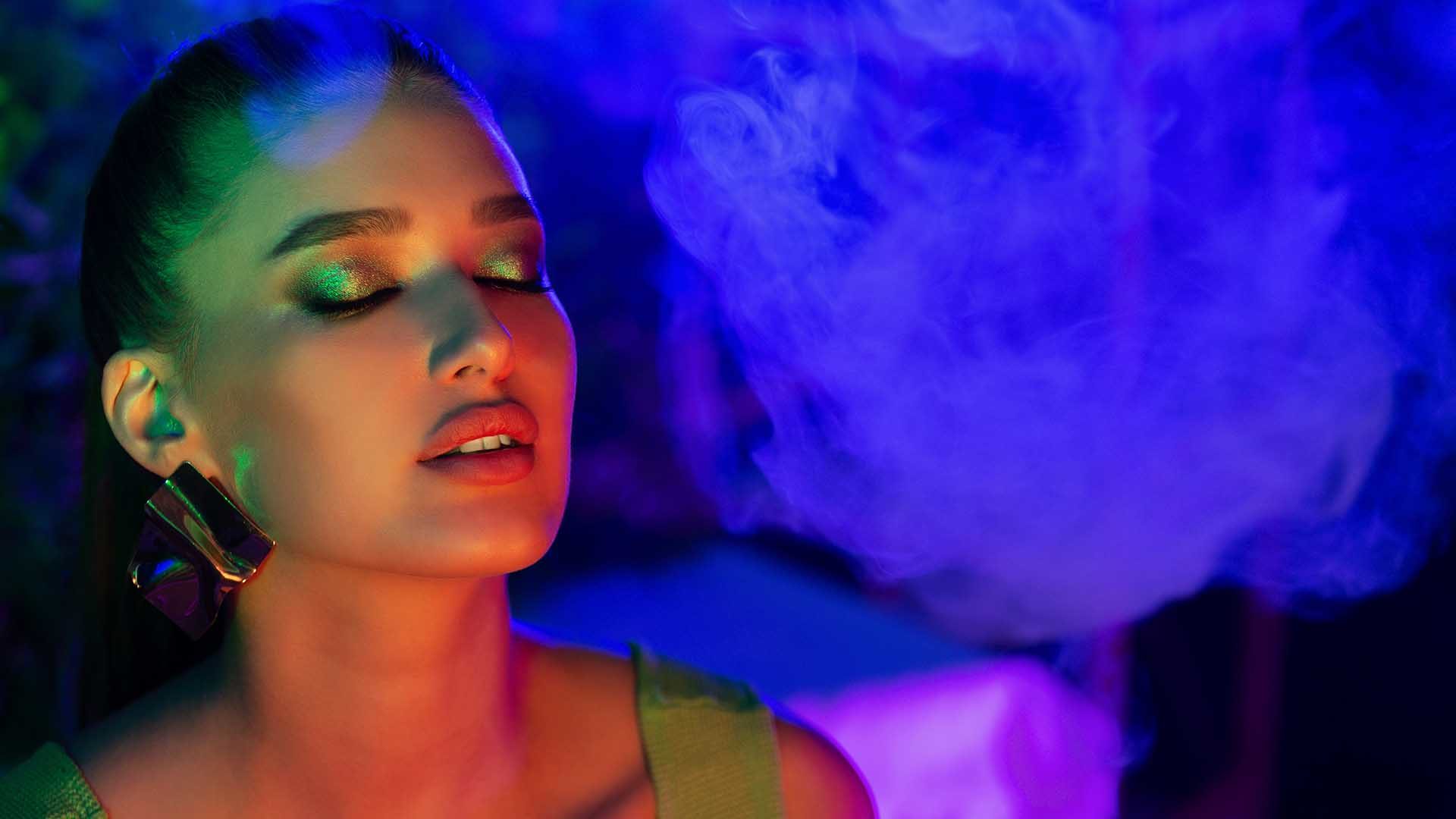 Young woman smoking vape in neon blue light, girl closed eyes, free space