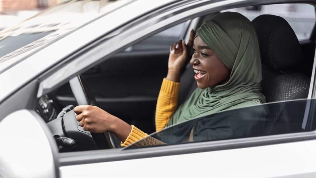 Driving Fun. Cheerful black muslim woman in hijab listening music and singing in car, excited african islamic lady enjoying drive in city, holding steering wheel and looking on the road, side view