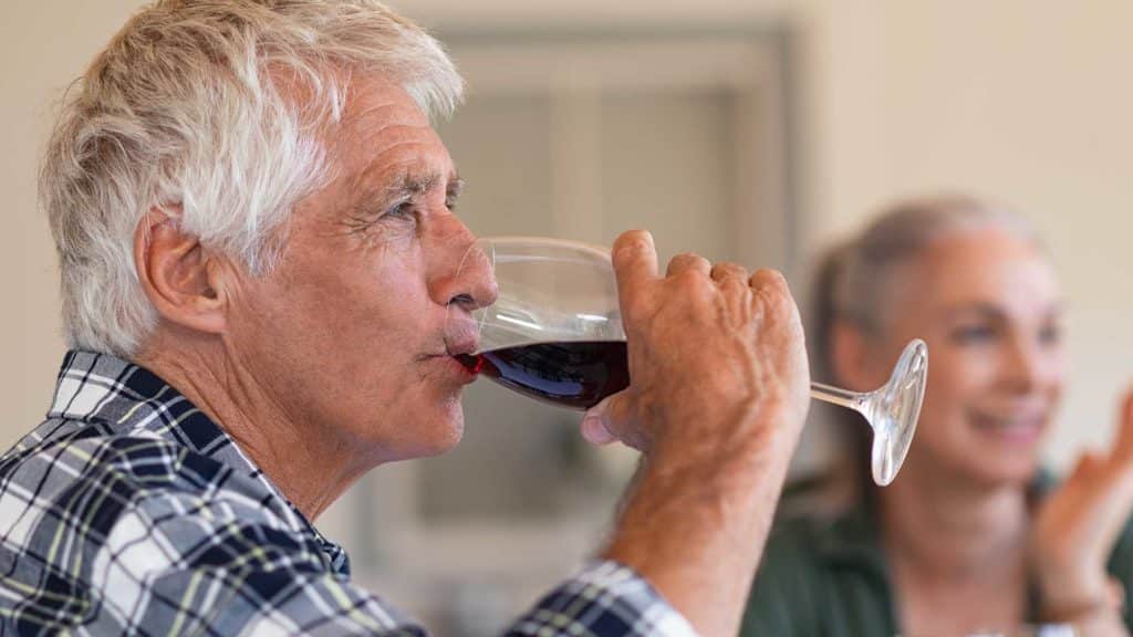 Happy senior man drinking a glass of red wine during lunch. Old man enjoying wine with friends in background. Closeup face of active and healthy senior man tasting wine.