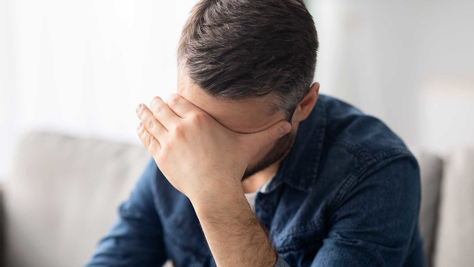 Upset middle-aged bearded man having headache, sitting on couch at home and touching his forehead, copy space. Closeup of frustrated man suffering from difficulties or depression, copy space