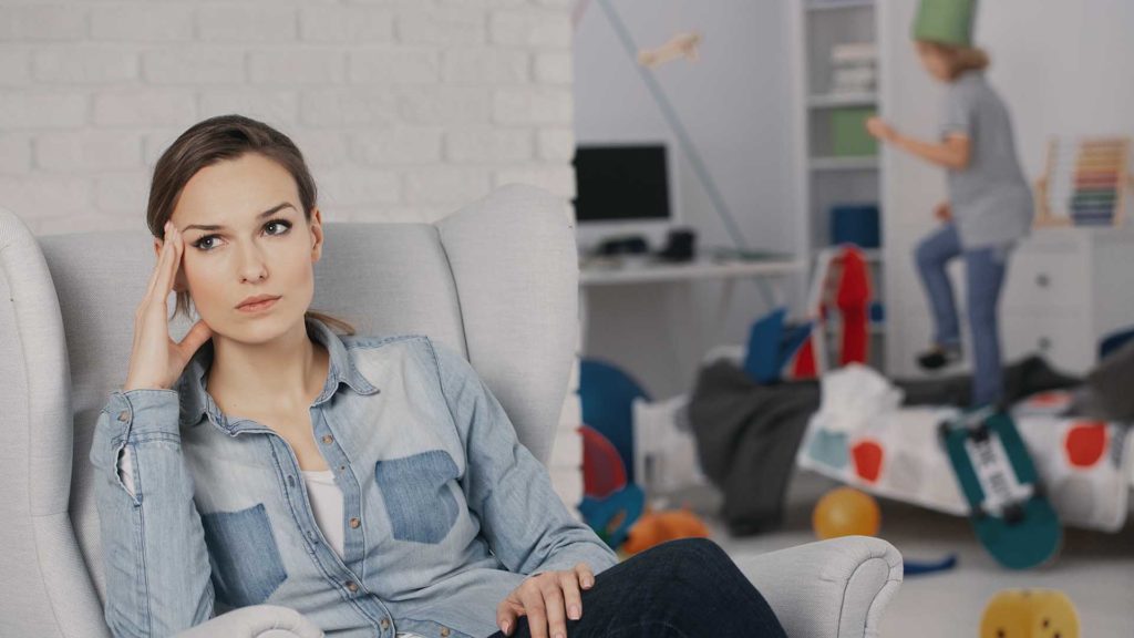 Worried mother relaxing on armchair while son with adhd making a mess