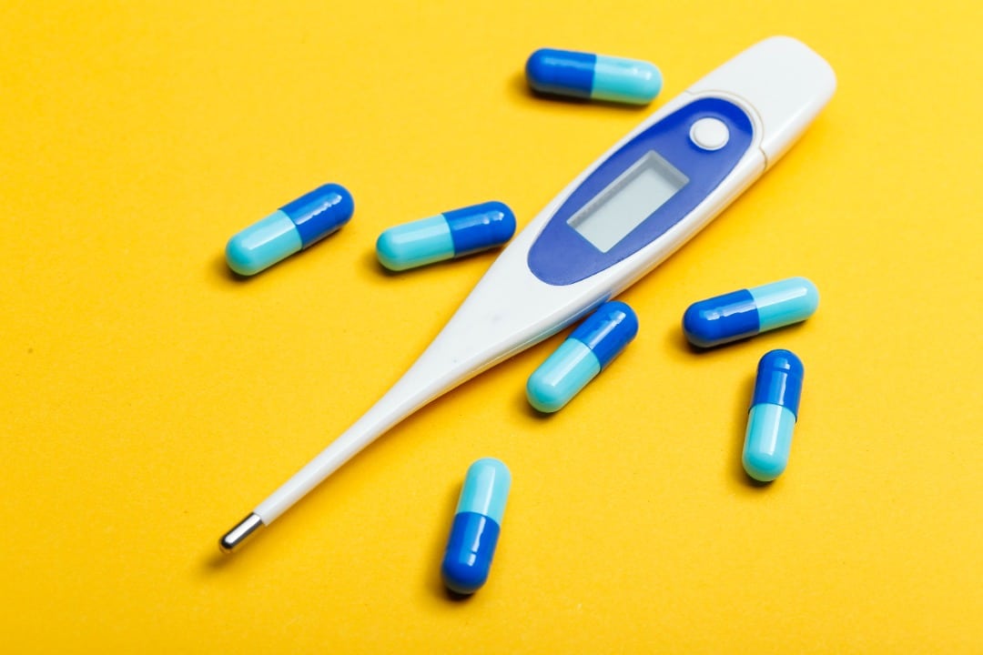 Electronic thermometer surrounded by blue pills on a yellow background.