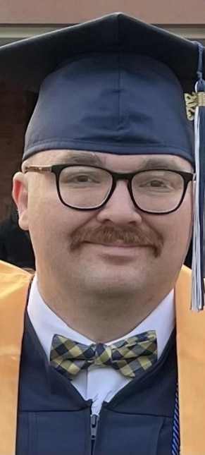 Seth is smiling at the camera. He has a mustache and wears glasses and a graduation cap and robe.